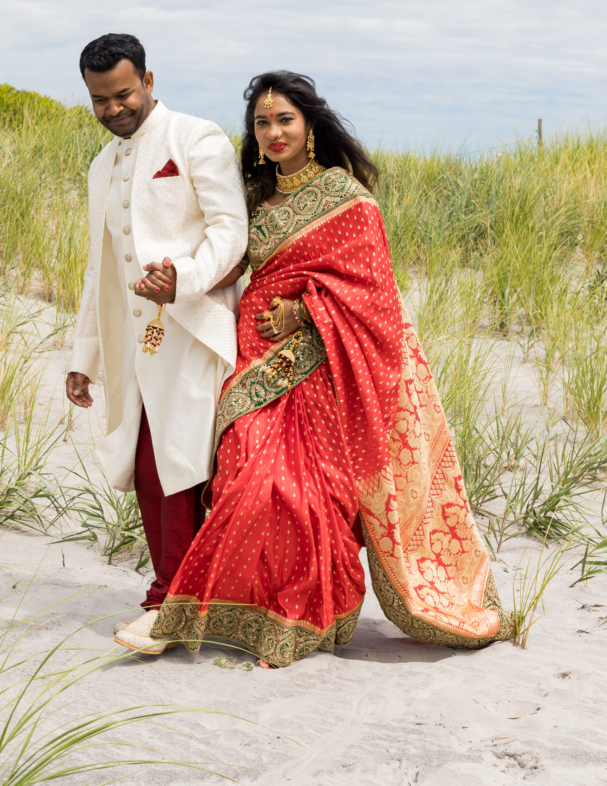 A South Asian bride in a red and gold embroidered saree standing with her groom in a white sherwani on a beach dune.